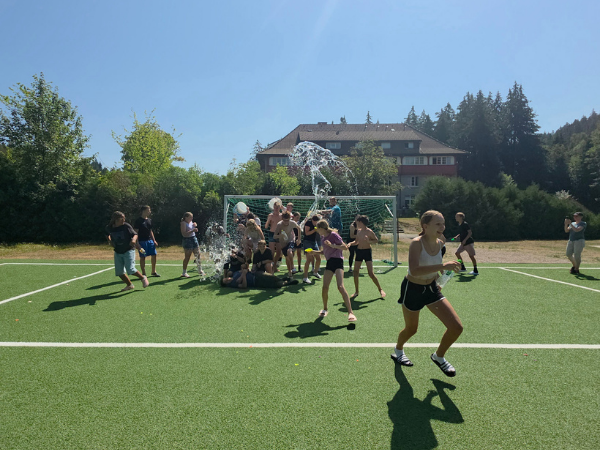 Schluchsee-Abkuehlung Eine Gruppe Jugendlicher steht im Sommer auf einem Kunstrasenplatz und kühlt sich mit Wasser ab, im Hintergrund ist ein Haus zu sehen, im Vordergrund läuft ein Mädchen mit Badeschlappen und Trinkflasche weg