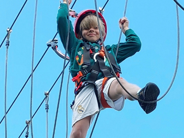Ein blonder Jugen mit rotem Kletterhelm klettert vor blauem Himmel in einem Kletterwald