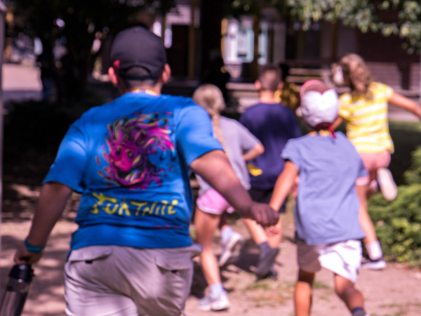 Sport-Camp Schönhagen Eine Gruppe Kinder rennt auf einem Gelände nach vorne, die Sonne scheint, ein Junge trägt ein blaues T-Shirt mit Fortnite