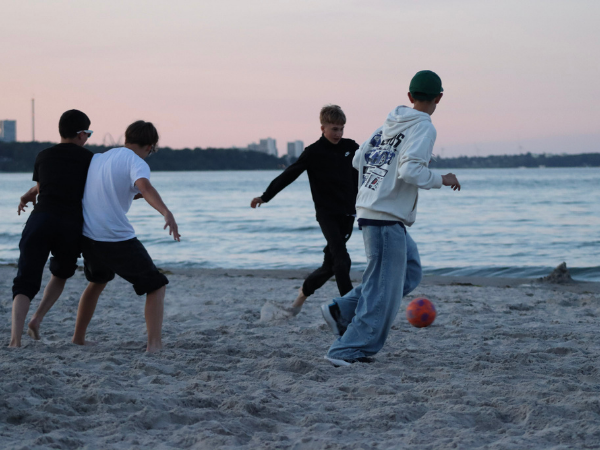 Vier Jungen spielen zum Sonnenuntergang Fußball am Strand im Hintergrund sind die Ostsee und ein paar Hochhäuser zu sehen
