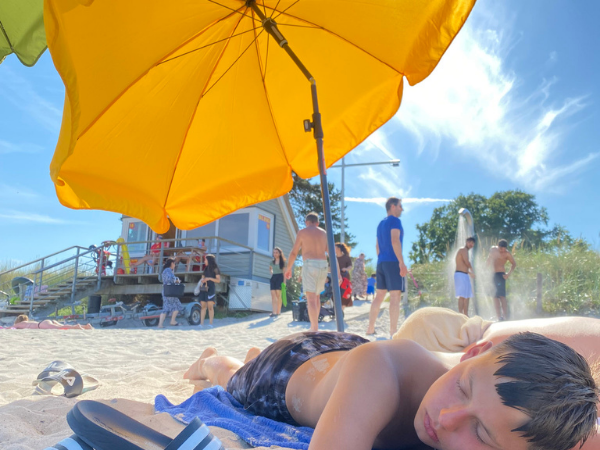 Zeltlager Scharbeutz Strand 2 Ein Junge liegt am Strand unter einem orangenen Sonnenschirm und schläft, im Hintergrund duschen zwei Jugendliche und eine Gruppe Menschen steht an einem Holzhaeuschen