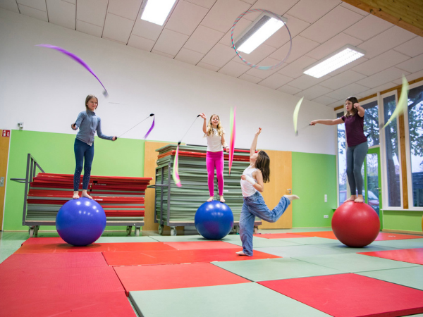 Vier Mädchen in einer Turnhalle, drei stehen auf Laufkugeln und haben Jongliertücher in der Hand ein Mädchen steht in der Mitte vor ihnen und wirft einen Ring in die Luft