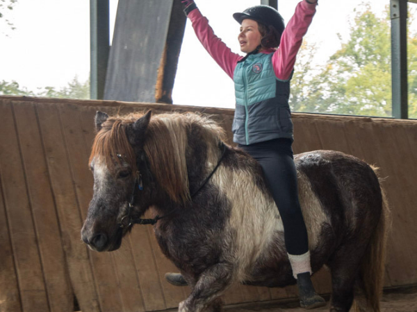 Naeve Herbst Mädchen sitzt auf einem Pony in einer Reithalle und reißt fröhlich die Arme nach oben