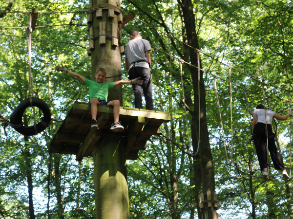 Mehrere Jugendliche klettern in einem Klettergarten in einem Wald, ein Junge sitzt auf einer Plattform und hat die Hände ausgestreckt, im Hintergrund hangeln zwei Jugendliche sich weiter