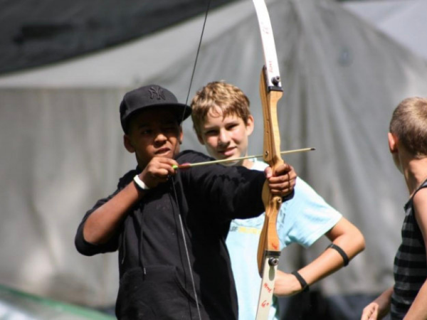 Camp Adventure Regen Ein June spannt einen Pfeil in einen Bogen ein, ein weiter Junge sieht im von hinten zu, im Hintergrund ist ein weißes Zelt zu sehen