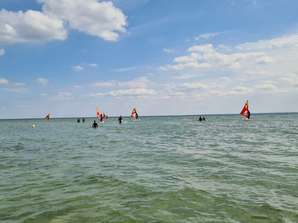 Am Horizont sind Windsurfer zu sehen, davor die blaue Ostsee und ein blauer Himmel mit weißen Wolken